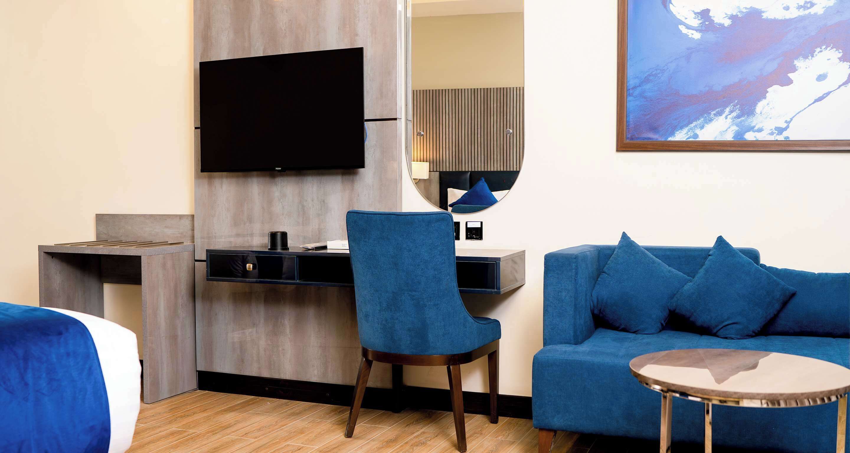 A modern hotel room with a flat-screen TV mounted on the wall, a desk with a chair, a blue sofa, and a small round table.