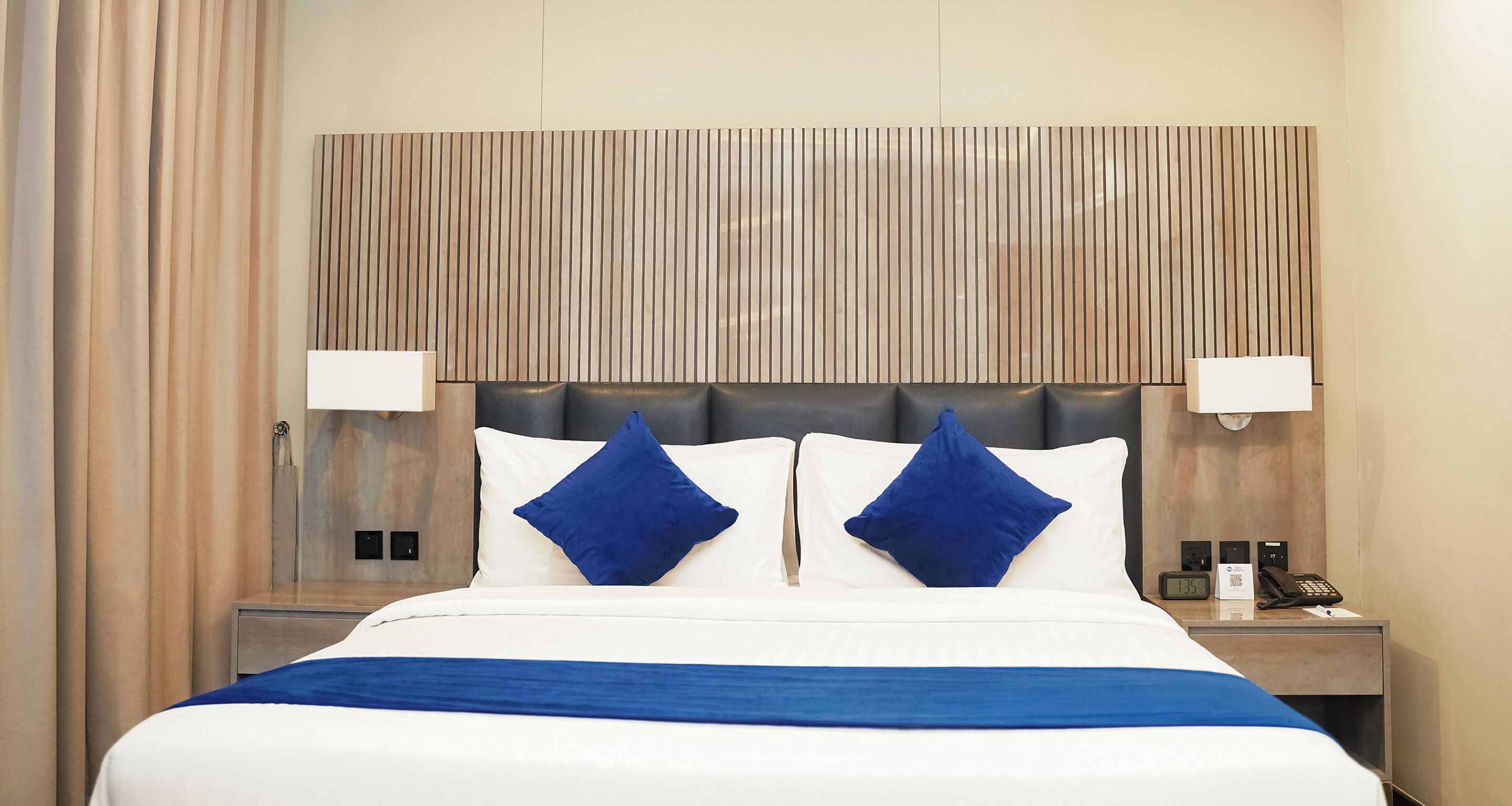 A hotel room with a bed, two bedside lamps, a nightstand with a phone and a digital clock, and a wall with a textured panel.