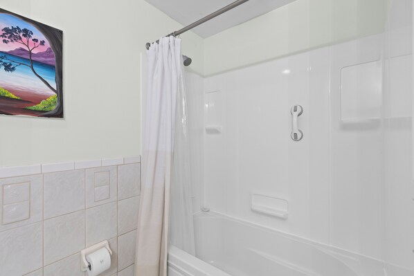 A bathroom with a shower, a toilet paper holder, and a painting of a landscape.