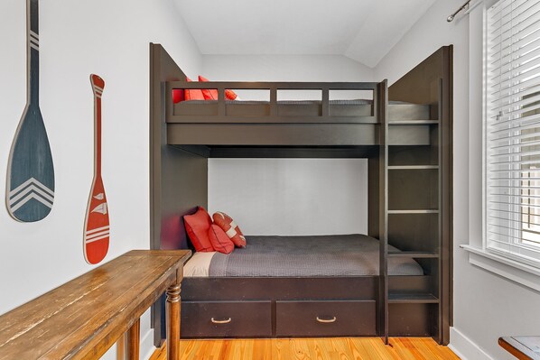 A bunk bed with a desk and drawers, a surfboard decoration, and a window with blinds.