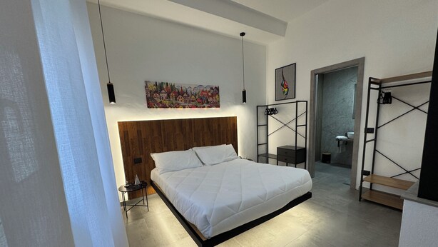 A modern bedroom with a large bed, a wooden headboard, a bedside table, a wall-mounted shelf, and a painting above the bed. - La Dimora del Centro - Agrigento Complete apartment in the heart of the City (Agrigento)