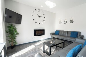 Ein modernes Wohnzimmer mit einer großen Wanduhr, einem Flachbildfernseher, einem Kamin, einem grauen Sofa mit blauen Kissen und einem Couchtisch.