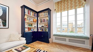 A cozy living room with a white sofa, a wooden coffee table with a book, a dark blue bookshelf, and a window with a view of buildings. - Charming 2 Bedroom 2 Bathroom, Notting Hill (London)