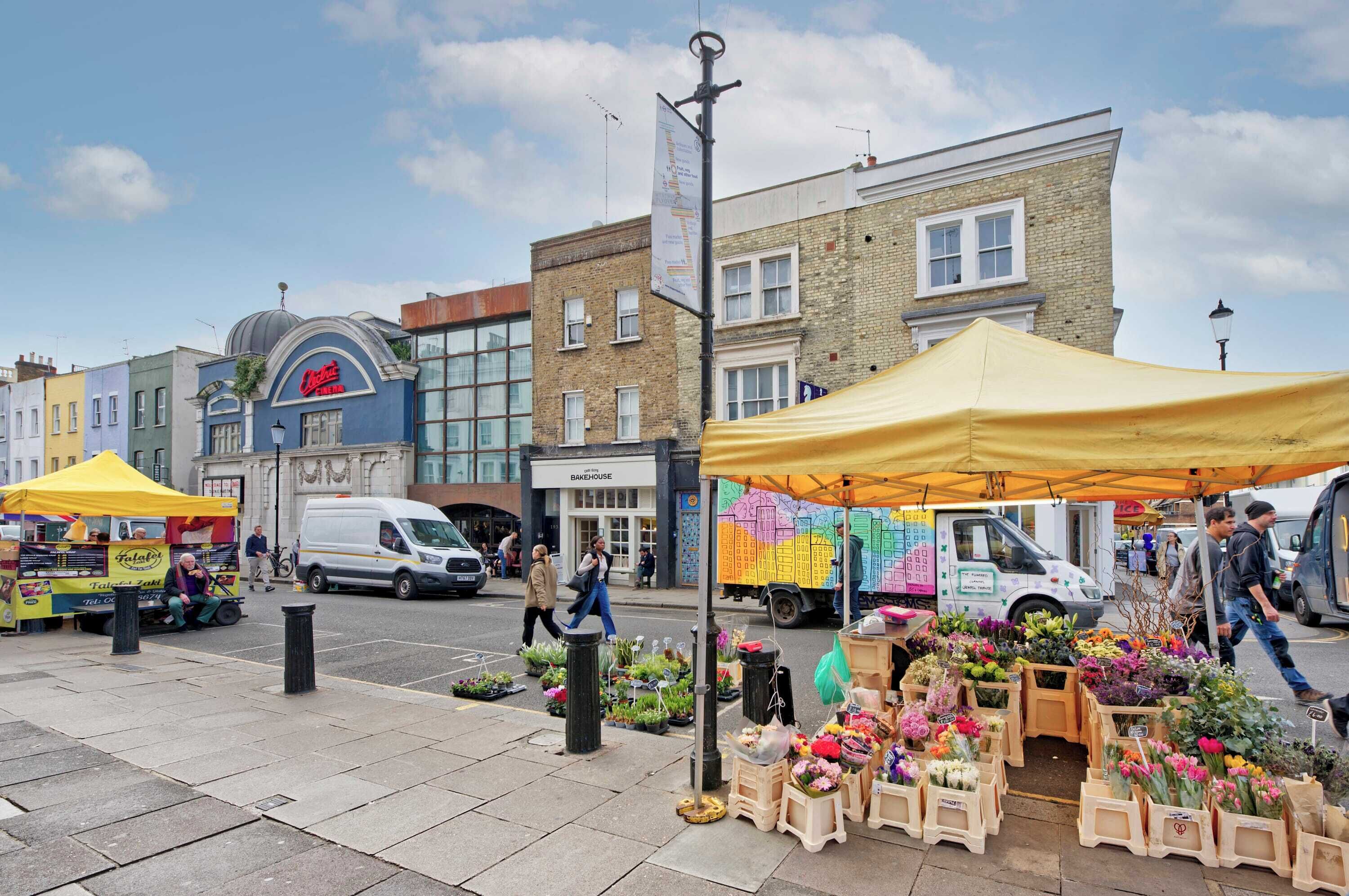 Un marché de rue avec des étals vendant des fleurs et de la nourriture, une boulangerie et un fourgon blanc garé sur le bord de la route.