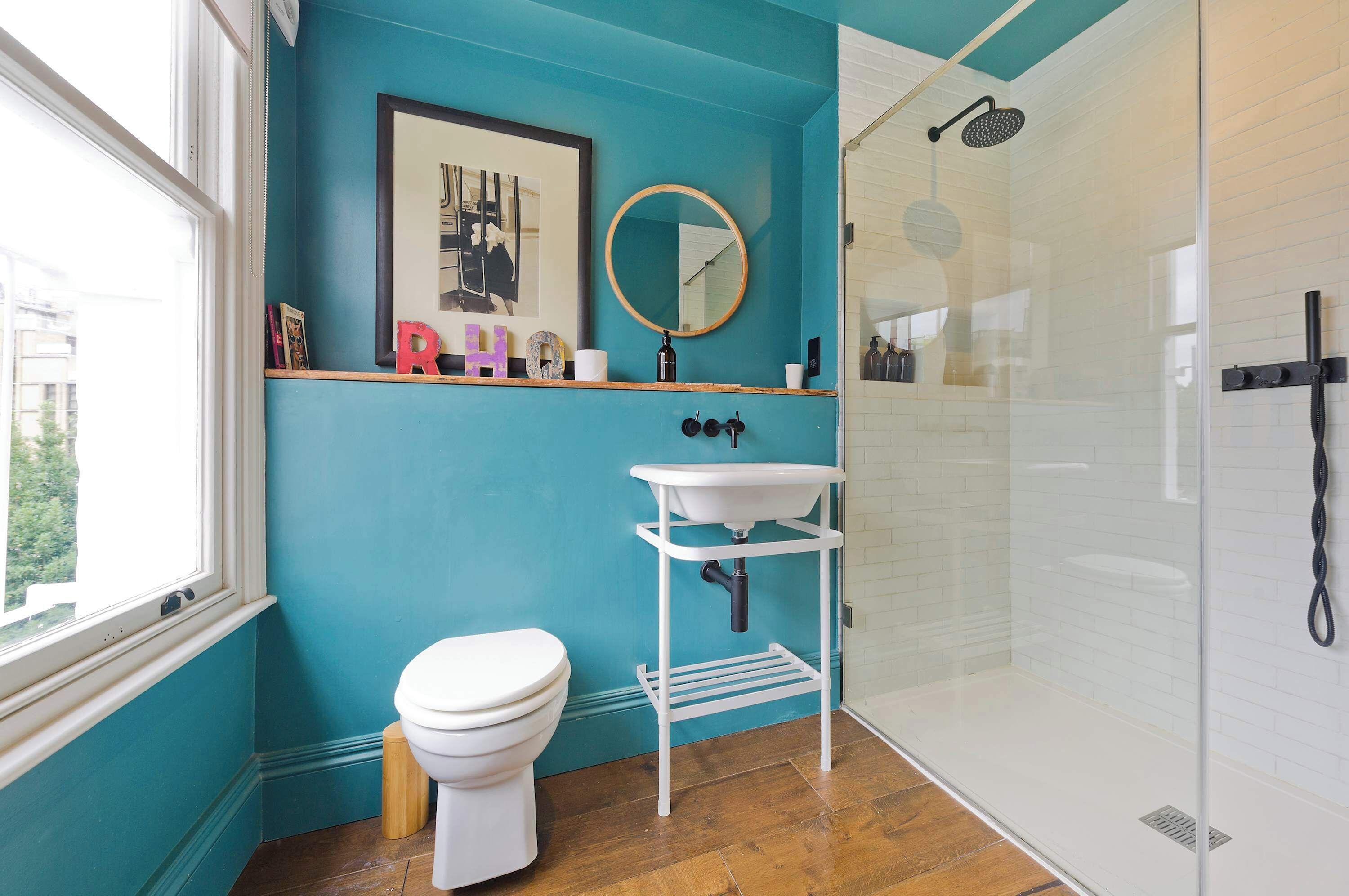 A bathroom with a white toilet, a white sink with a mirror, a shower, and a teal wall.