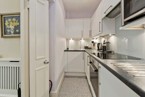 A narrow kitchen with white cabinets, a black countertop, and a microwave. - Studio Flat With Garden Access, Holland Park (London)