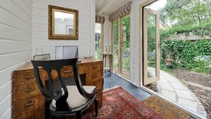 A room with a wooden desk, a chair, a mirror, and a large window opening to a garden.