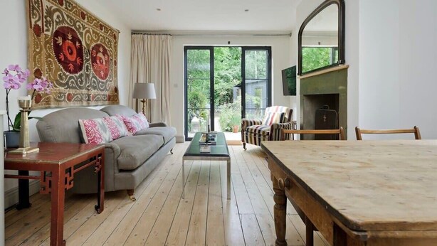 A living room with a sofa, a coffee table, a dining table, a large tapestry, a mirror, and a view of greenery outside.