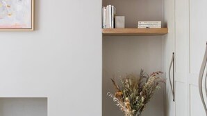 Et minimalistisk rum med en hvid væg, et indrammet abstrakt maleri, en træbogreol og en glaskrukke med tørrede blomster på en piedestal.