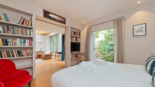 A modern hotel room with a large bed, a red armchair, and bookshelves filled with books. - 5 Bedroom House, Maida Vale (London)