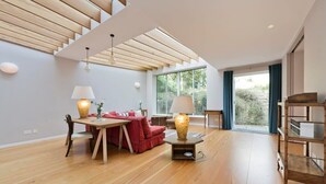 A spacious living room with a wooden floor, a dining area with a table and chairs, a red sofa, and a large window providing a view of greenery outside. - 5 Bedroom House, Maida Vale (London)