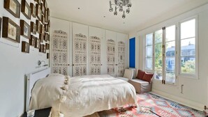 A bedroom with a large bed, a patterned rug, a wall of framed pictures, and a window with a view of buildings. - Stylish 5 Bed House in St. John's Wood/maida Vale (London)
