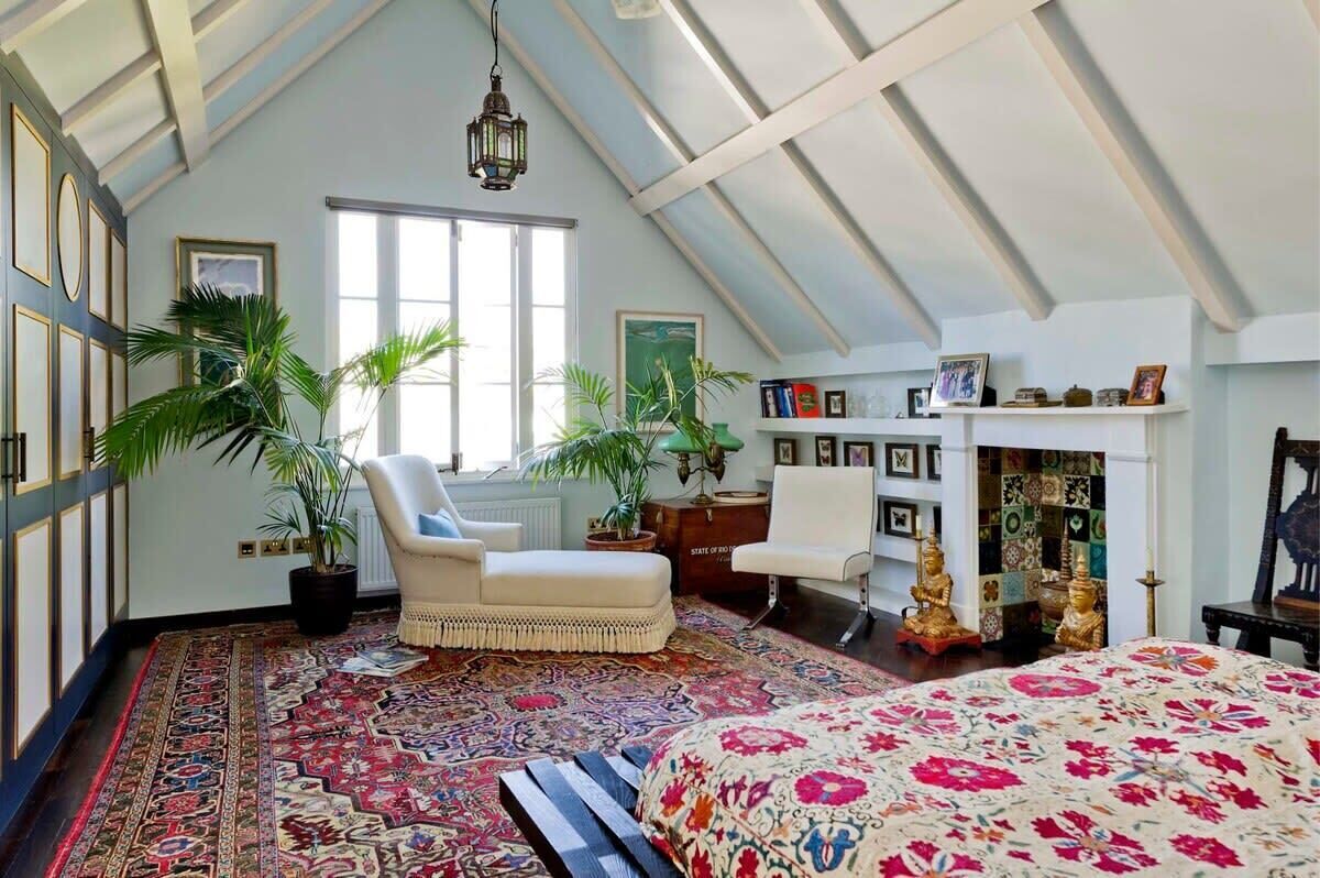 A room with a sloped ceiling, a large bed with a floral bedspread, a chair, a small table, and two potted plants.