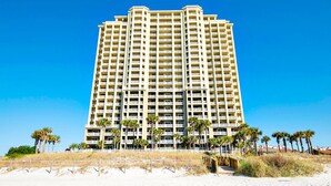 Un hotel alto de varios pisos con balcones, situado cerca de una playa de arena con palmeras.