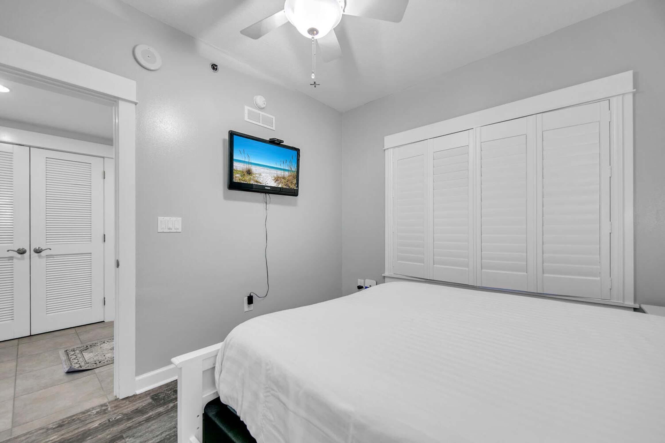 A bedroom with a bed, a ceiling fan, a TV mounted on the wall, and a set of white closets.