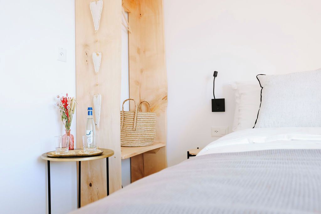 Een slaapkamer met een bed, een nachtkastje met een waterfles, een geweven mand en een wandcontactdoos.