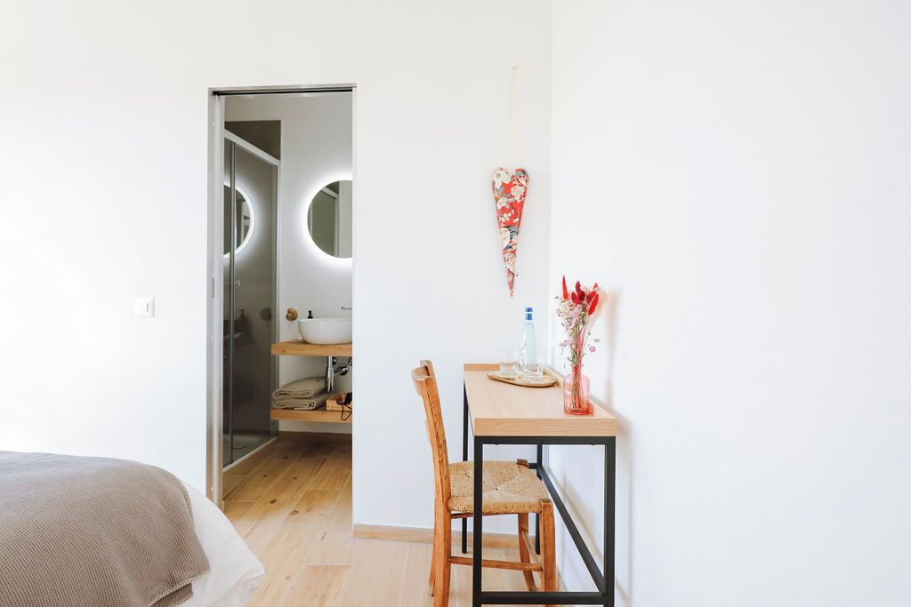 A bedroom with a bed, a wooden chair, a small table with a vase, and a bathroom with a circular mirror.