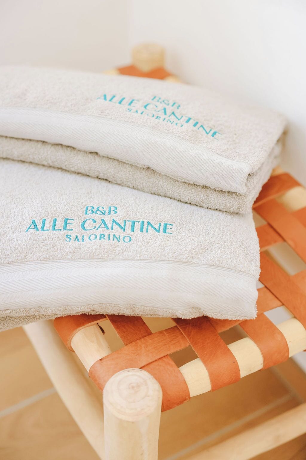 Une pile de serviettes blanches sur lesquelles est imprimé le texte « B&B ALLE CANTINE SALORINO », posée sur une chaise en bois dont l’assise est en osier tressé.