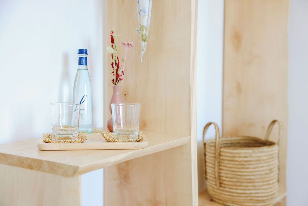 A wooden shelf with a clear glass bottle, a clear glass, and a pink vase with dried flowers.