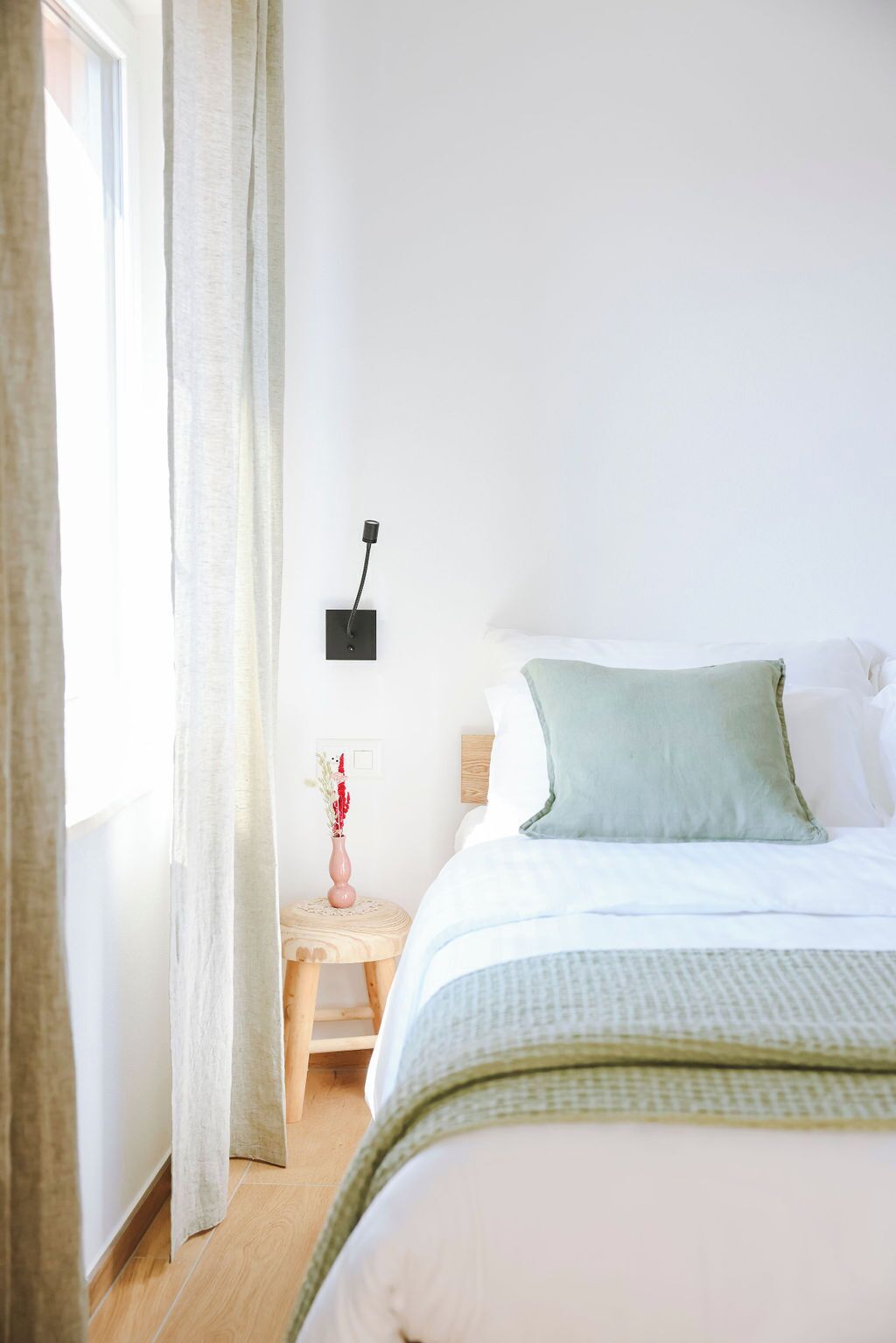 Quarto com cama, mesa de cabeceira, abajur e janela com cortinas.