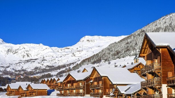 Une rangée de cabanes en bois aux toits enneigés, se dressant sur un fond de montagnes recouvertes de neige et d’un ciel bleu clair.