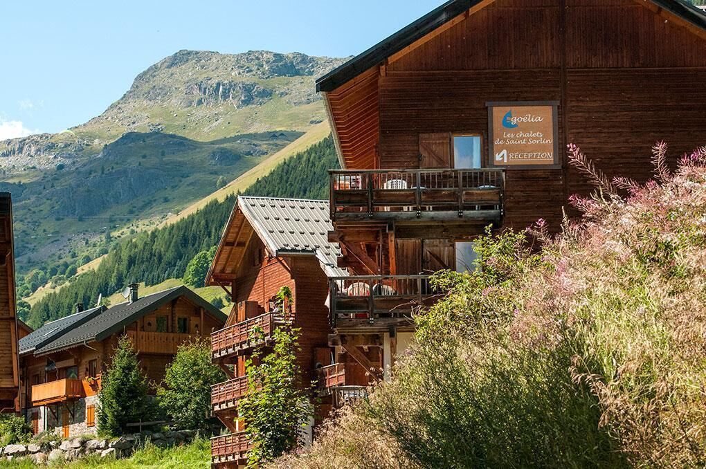 A wooden chalet with a balcony, surrounded by mountains and greenery.
