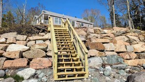 A wooden staircase leading to a house with a stone foundation, surrounded by trees and rocks. - Beachfront House w/Fire Pits & Ocean Views, (Ellsworth)