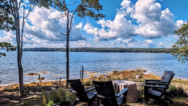 A lakeside seating area with chairs and a table, surrounded by trees and a view of the lake. - Beachfront House w/Fire Pits & Ocean Views, (Ellsworth)