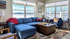 A living room with a sectional sofa, a coffee table, and large windows with a view of a snowy landscape. - Beachfront House w/Fire Pits & Ocean Views, (Ellsworth)