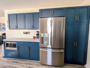 A kitchen with blue cabinets, a stainless steel refrigerator, a coffee maker, and a microwave. - Beachfront House w/ Fire Pits & Private Access (Ellsworth)