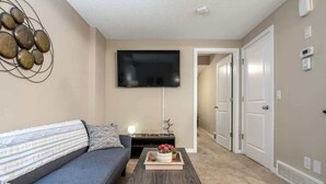 A living room with a gray sofa, a coffee table with a tray of flowers, a flat-screen TV mounted on the wall, and a hallway leading to a door. - The Grande 1BD 1BA - Full Kitchen Guest Suite (Calgary)