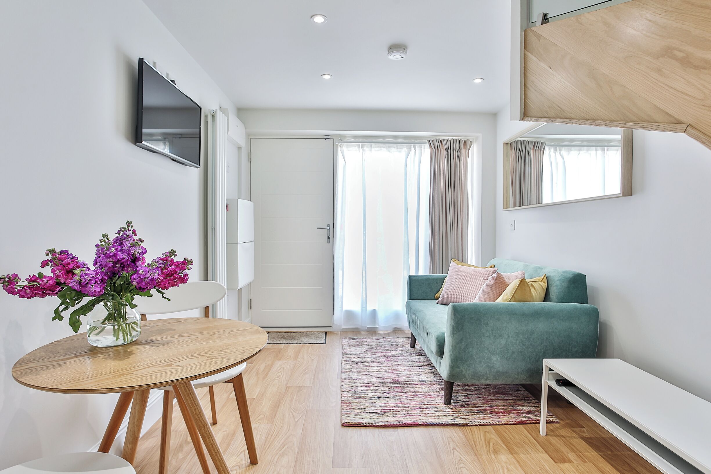 A modern living room with a green sofa, a wooden dining table with chairs, and a TV mounted on the wall.
