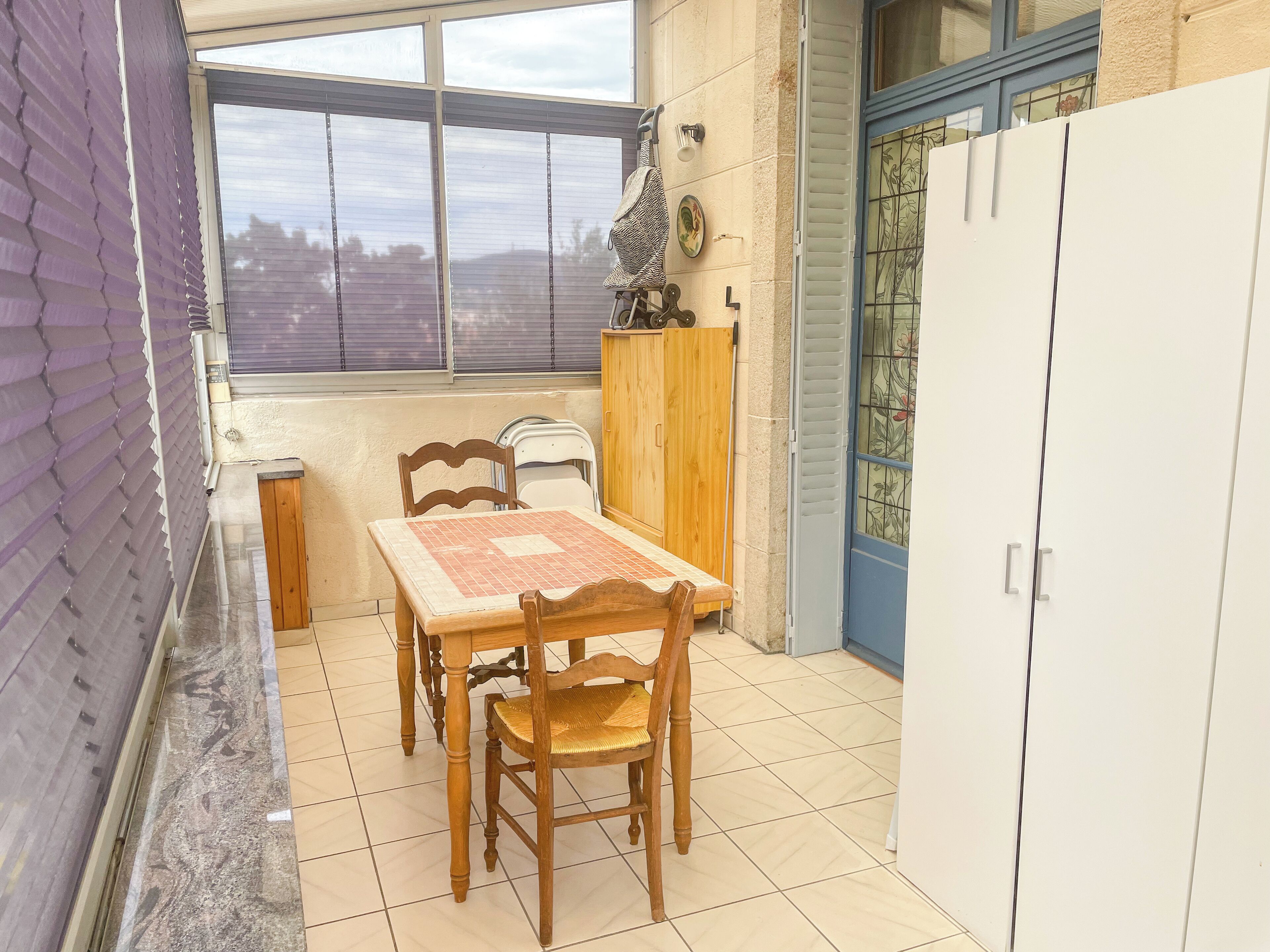 Un pequeño comedor con una mesa y sillas de madera, una ventana con persianas, un armario y una puerta con un panel de vidrio decorativo.