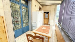 A small, enclosed patio area with a tiled floor, a wooden table, a wicker chair, a blue-framed door with decorative glass, and a white refrigerator. - L'Arpège du Puy - House* ParkingView * Family* 10min walk from Center (Le Puy-en-Velay)