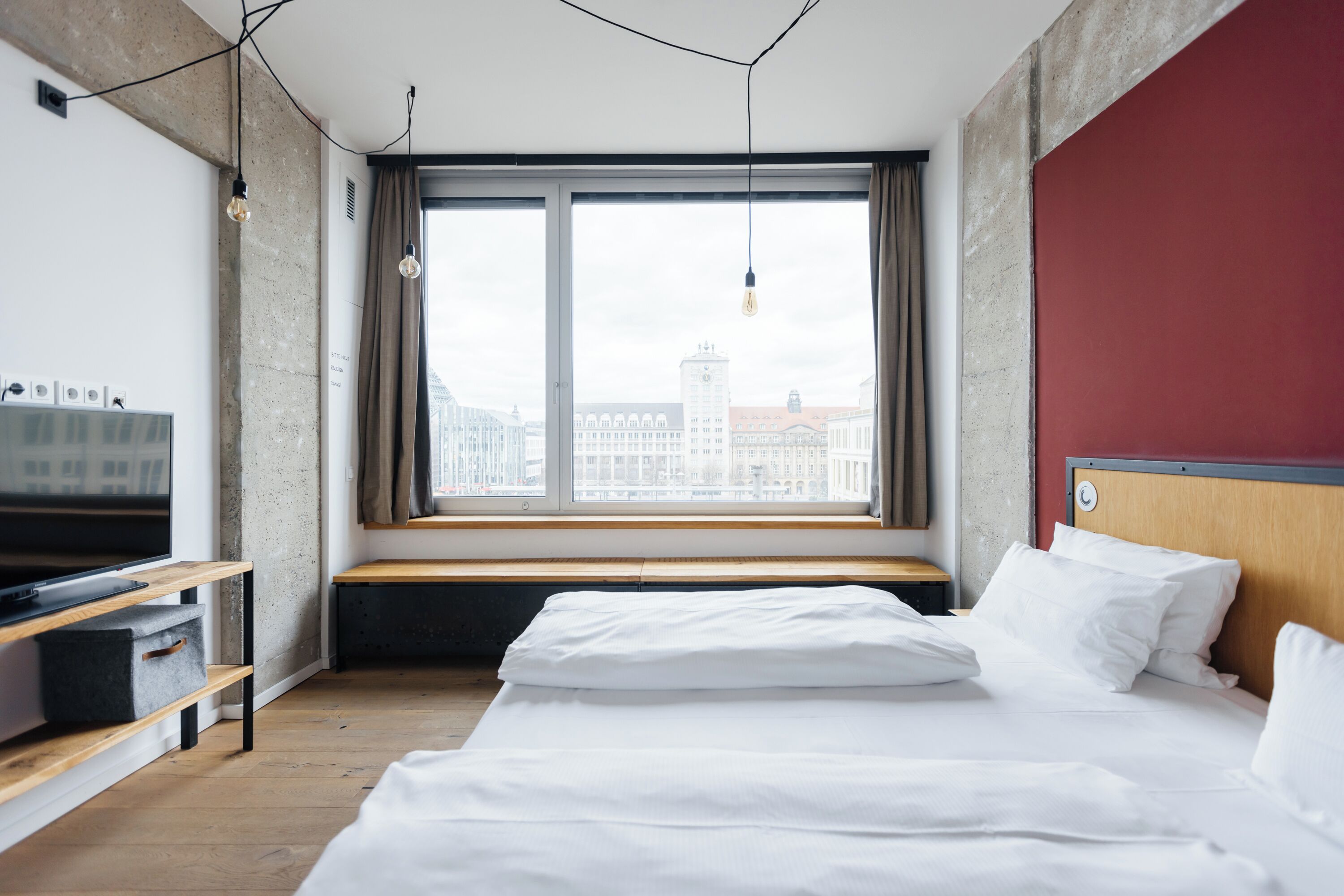 A modern hotel room with a large window, a bed, a TV, and a wooden bench.