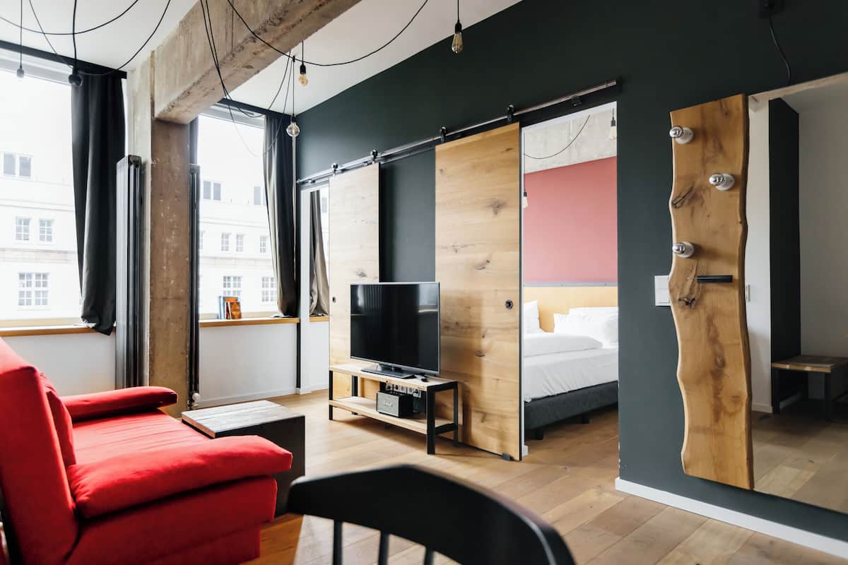 Una habitación moderna con un sofá rojo, un televisor sobre un mueble de madera, una cama con pared rosa y una puerta grande de madera.