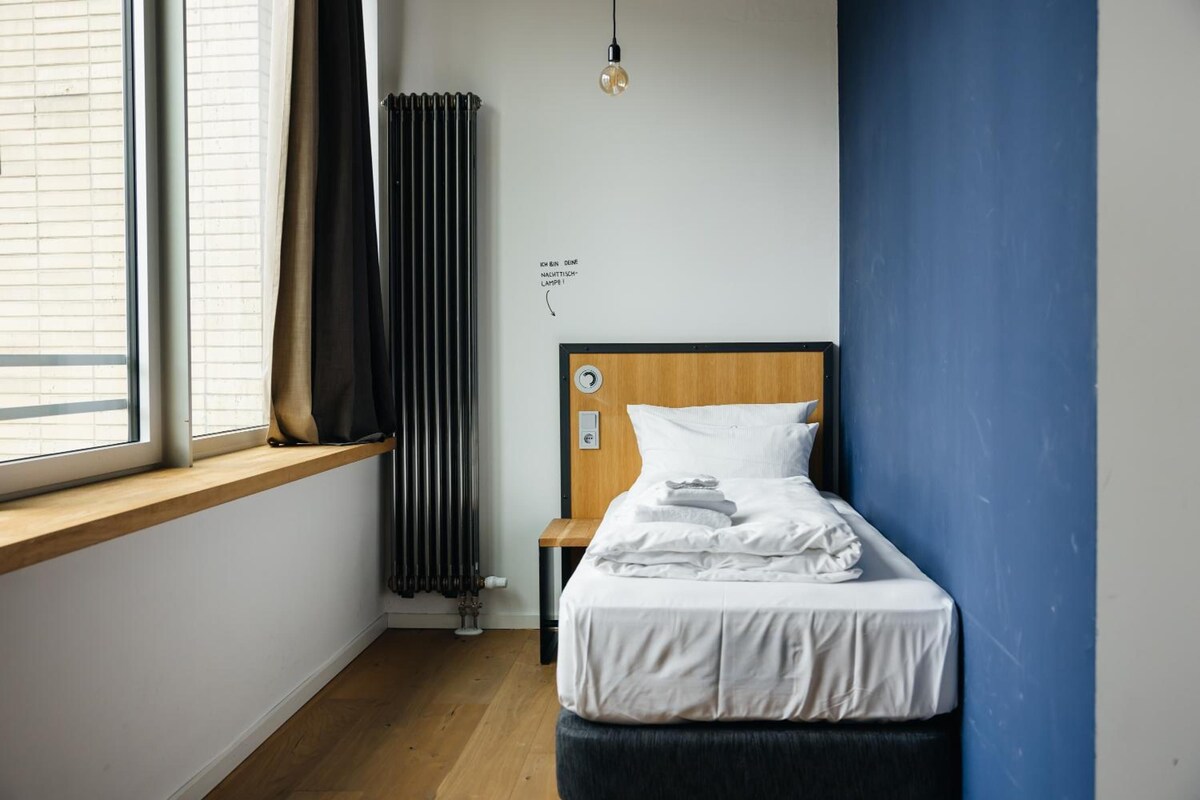 Un dormitorio con cabecera de madera, una ventana con persianas, un radiador y una mesita de noche con lámpara.