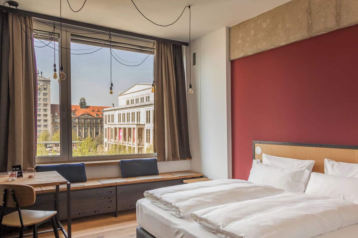 A hotel room with a bed, a desk, and a view of buildings through the window.