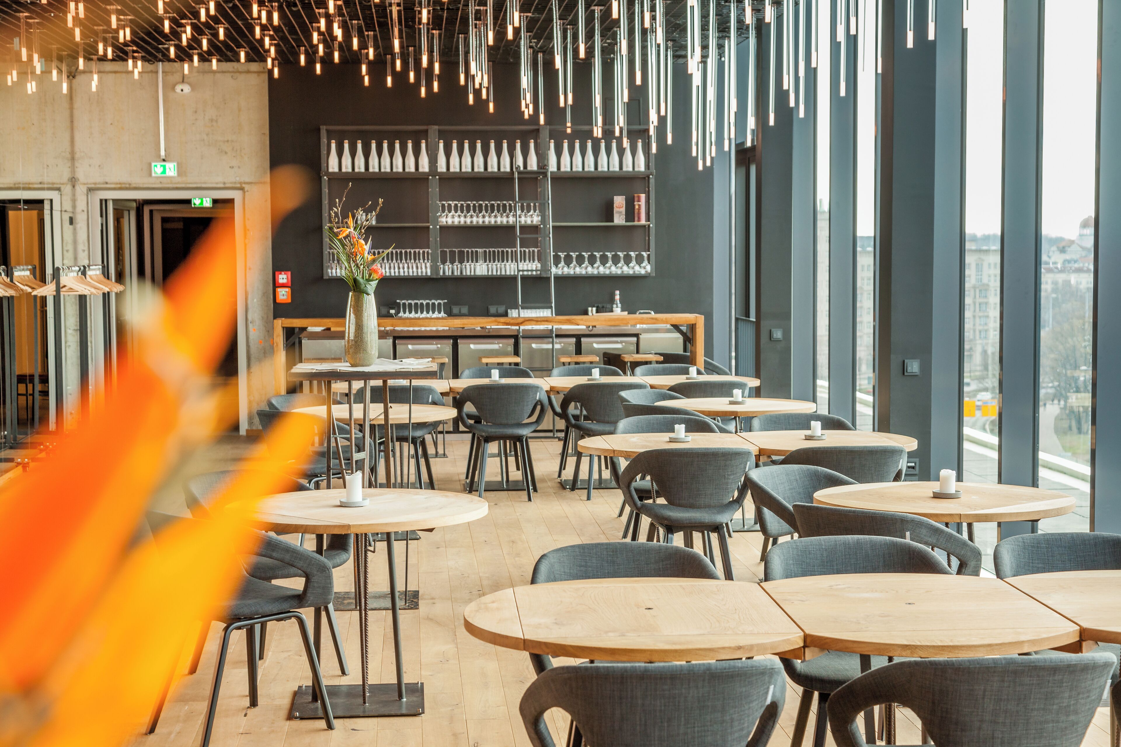A modern cafe with round tables and chairs, a large window with a view, and a shelf with glassware.