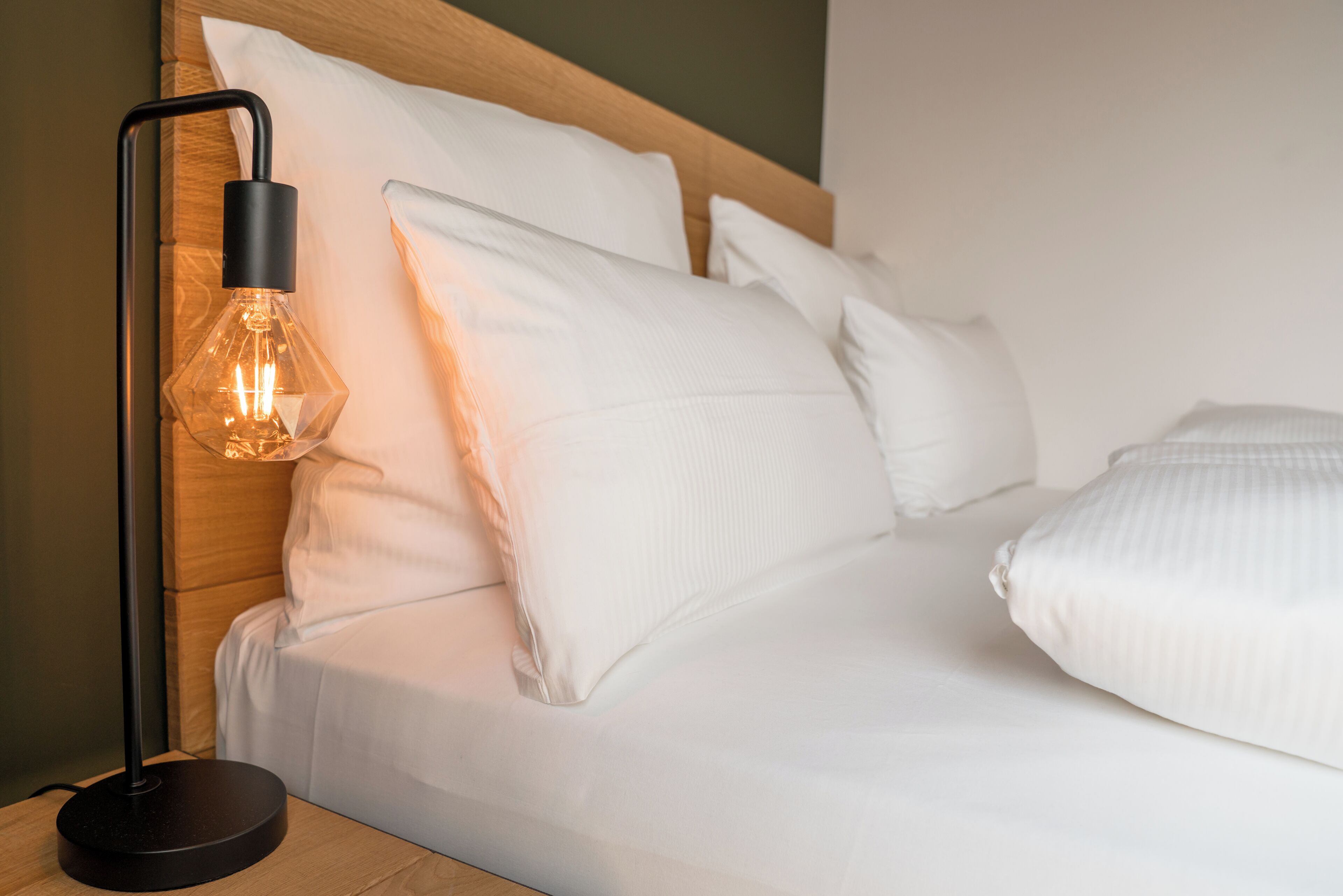 Una cama bien tendida con ropa blanca y almohadas, una lámpara de pie negra moderna con pantalla de vidrio transparente, y un cabecero de madera.