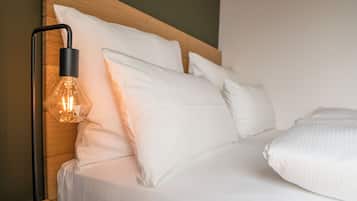 Una cama bien tendida con ropa blanca y almohadas, una lámpara de pie negra moderna con pantalla de vidrio transparente, y un cabecero de madera.