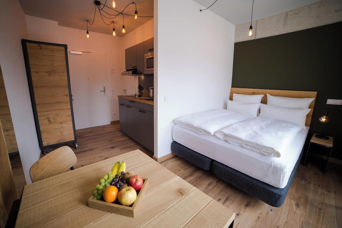 A modern hotel room with a wooden floor, a bed with white linens, a small kitchen area, and a dining table with fruit.