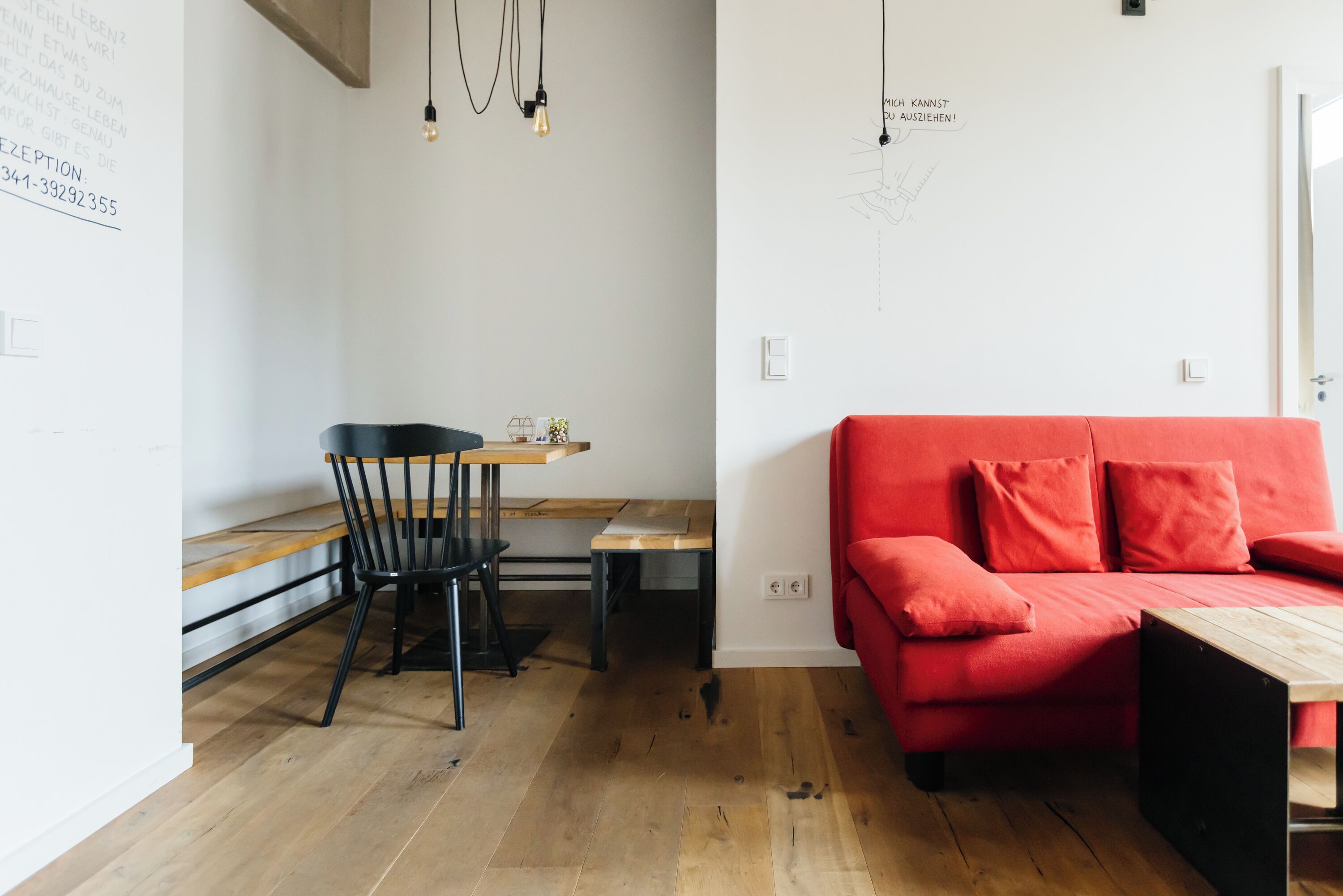 A modern room with a red sofa, wooden flooring, a dining table with chairs, and a wall with a light fixture and a sign.