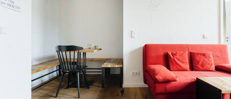 Una habitación moderna con sofá rojo, suelo de madera, una mesa de comedor con sillas, y una pared con lámpara y un cartel.