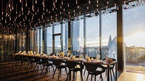 Une salle à manger spacieuse avec une longue table dressée pour le repas, de grandes fenêtres offrant une vue sur la ville et des luminaires suspendus.
