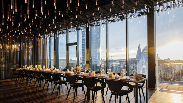 Une salle à manger spacieuse avec une longue table dressée pour le repas, de grandes fenêtres offrant une vue sur la ville et des luminaires suspendus.