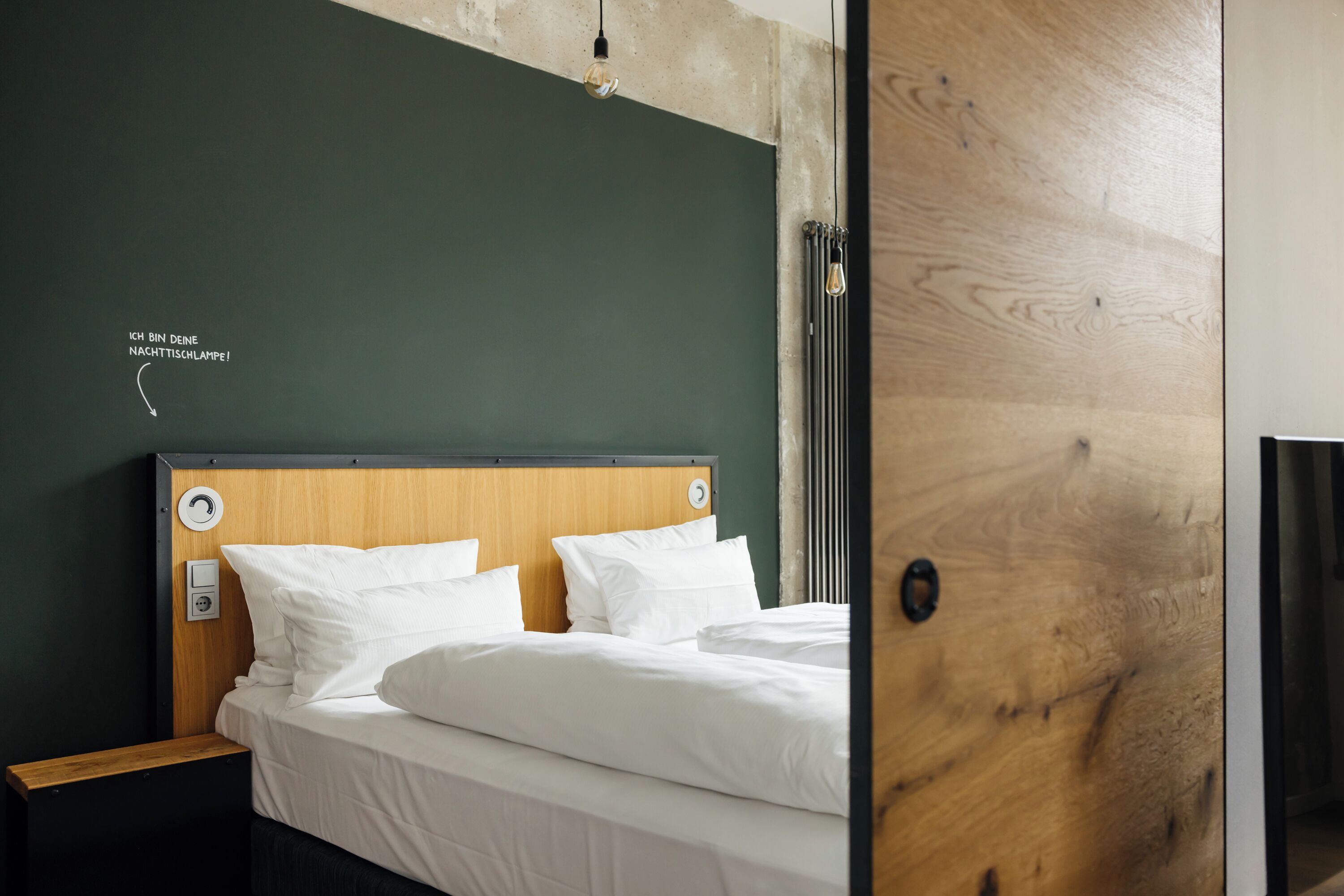 Una cama bien hecha con sábanas blancas, cabecera de madera y lámpara fijada a la pared.