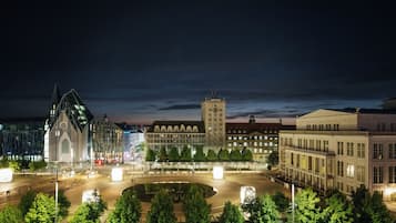 Ein nächtliches Stadtbild mit einem markanten Kirchturm, einem großen Gebäude mit einem Uhrturm und einer Baum-umgebenen Kreisverkehr.