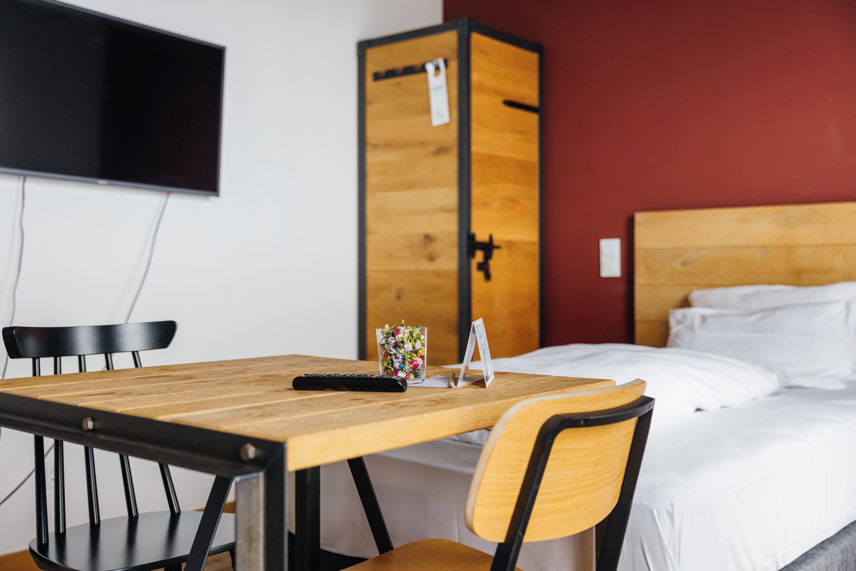 Habitación de hotel con una mesa de madera, dos sillas, una cama con ropa de cama blanca, un televisor montado en la pared y un armario de madera.