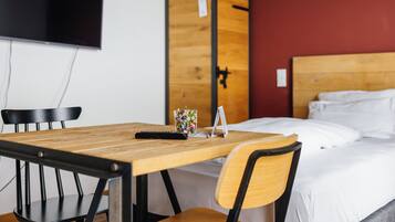 A hotel room with a wooden table, two chairs, a bed with white bedding, a TV mounted on the wall, and a wooden wardrobe.